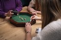 Vier mensen spelen een dobbelspel rond een achthoekige bak met groen vilt. Twee dobbelstenen liggen in de bak.