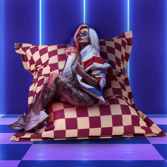 Vrouw met blonde kuif en tattoo's zittend op paars-crèmekleurige dambord zitzak met neonlicht.