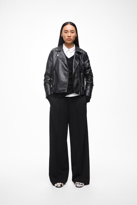 Model in zwart leren jack, wit overhemd en zwarte wijde broek.
