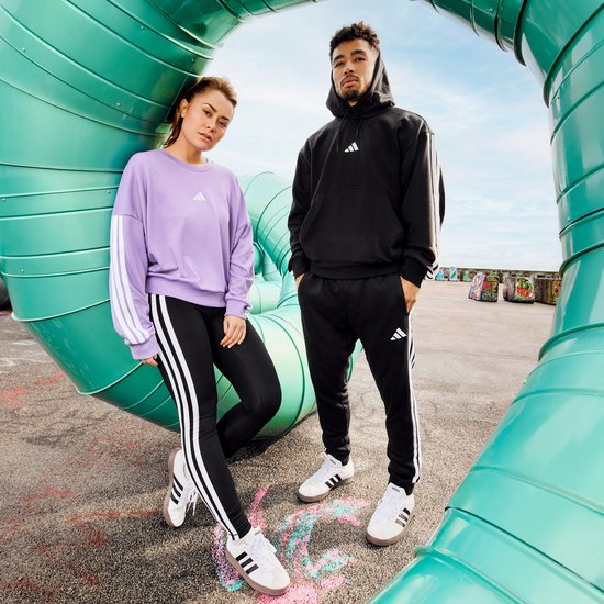 Een man en vrouw poseren buiten. De vrouw draagt een paarse trui en zwarte legging, de man een zwarte hoodie en joggingbroek.