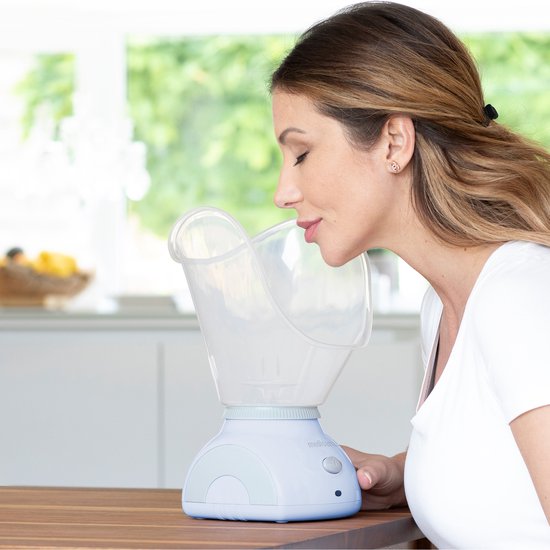 Vrouw met een witte top inhaleert stoom uit een witte Medisana gezichtssauna met transparante kap.