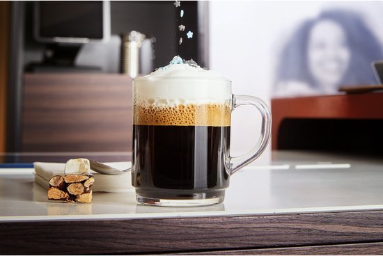 Een glas koffie met schuim en decoraties, met een stukje gebak ernaast.