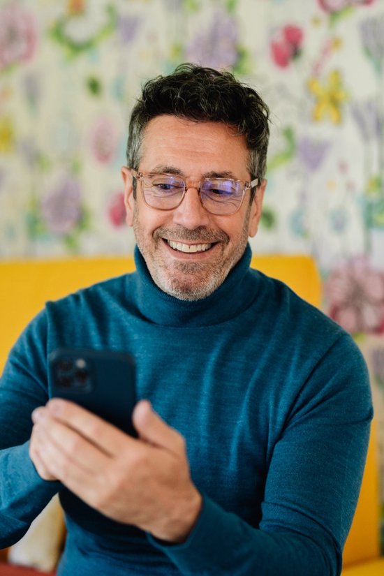 Homme barbu avec un col roulé gris, regardant un smartphone. Porte des lunettes.
