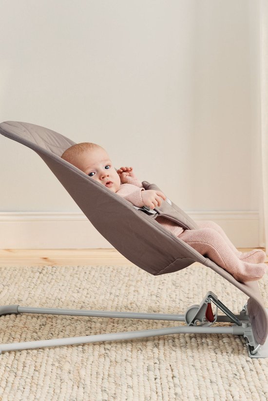 Baby met blauwe ogen in zandgrijs wipstoeltje, draagt roze gebreide kleding.