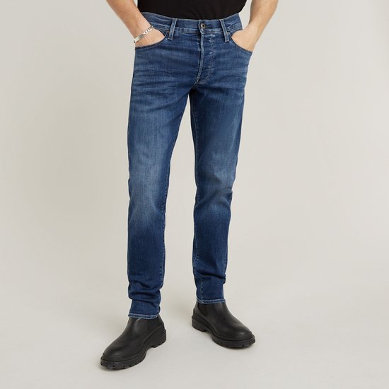 Vooraanzicht van de benen van een man in een donkerblauwe slim fit jeans met lichte wassing en zwarte schoenen.