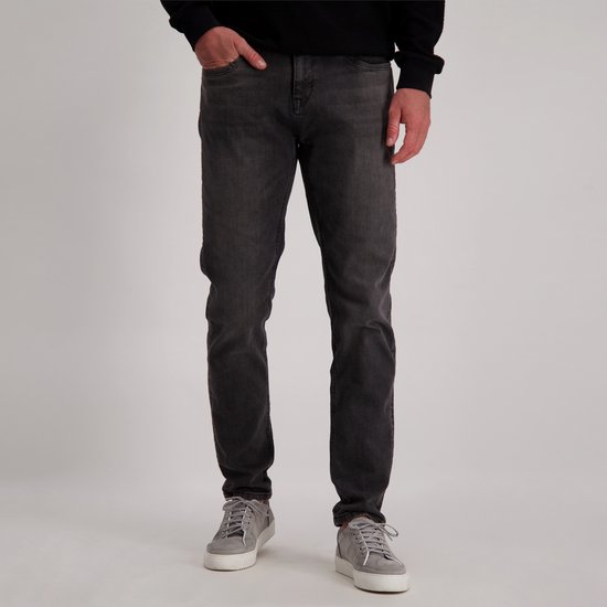 Man met donkere spijkerbroek en grijze sneakers, hand in broekzak.