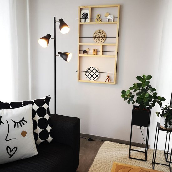 Zwarte staande lamp met drie spots in een woonkamer naast een zwarte bank met grafische kussens.