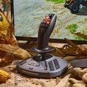 Zwarte Thrustmaster joystick met oranje knoppen op een grijze basis, omringd door herfstbladeren en maïskolven.