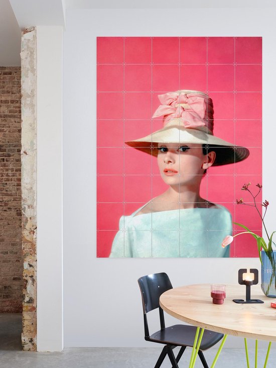 Audrey Hepburn portant un grand chapeau, un nœud rose et un haut bleu pâle, sur fond rose, près d'une table ronde et de chaises.
