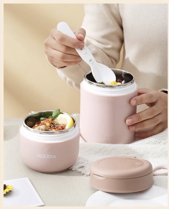 Twee roze lunchpots op tafel, gevuld met voedsel. Een hand houdt een lepel vast boven een pot.