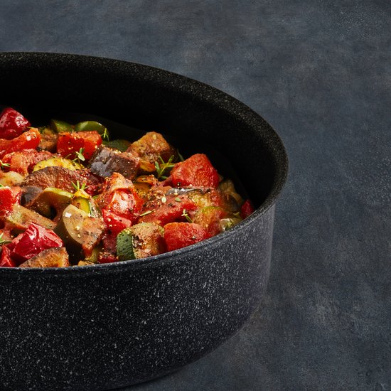Gestoofde groenten, waaronder courgette, aubergine en tomaat, in een zwarte hapjespan met kruiden.