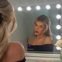 Vrouw met blond haar en rode lippen kijkt in een spiegel met verlichting.
