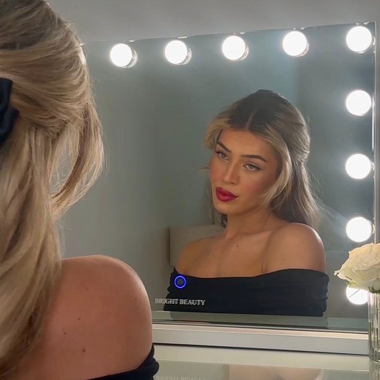 Vrouw met blond haar en rode lippen kijkt in een spiegel met verlichting.