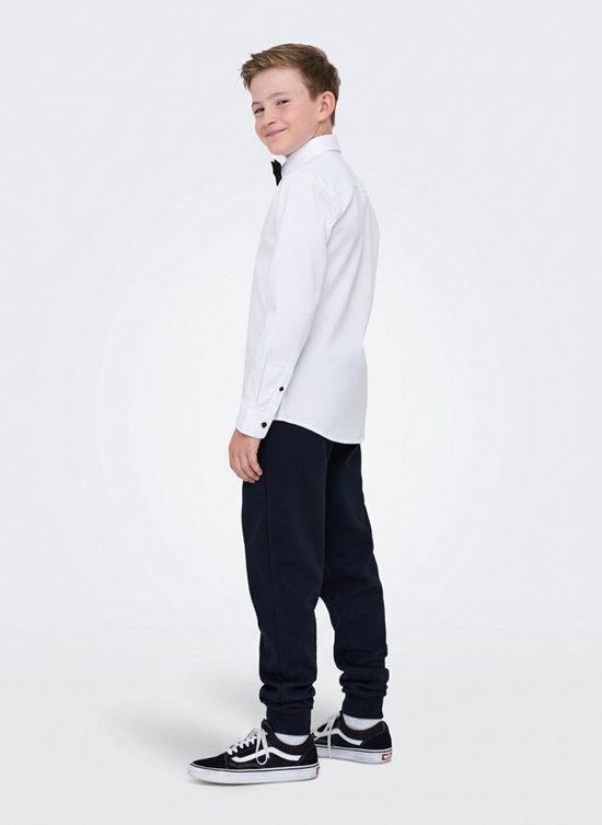 Jongen in wit overhemd met zwarte vlinderdas en zwarte knopen, staand opzij met zwarte broek en sneakers.