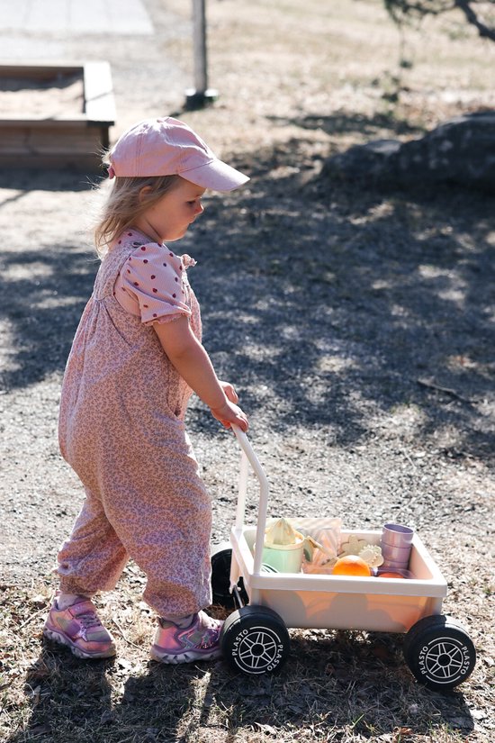 Een meisje in een roze tuinbroek duwt een zandkleurige Plasto kinderwagen met speelgoed en fruit.