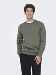 Een man met krullend haar draagt een mosgroene sweater met ronde hals en zwarte broek, met zijn handen in zijn zakken.