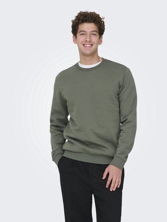 Een man met krullend haar draagt een mosgroene sweater met ronde hals en zwarte broek, met zijn handen in zijn zakken.