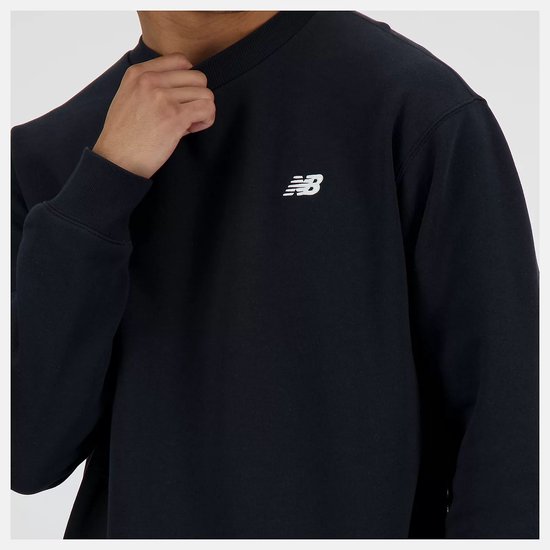 Close-up van een zwarte crewneck trui met een wit New Balance logo op de borst.