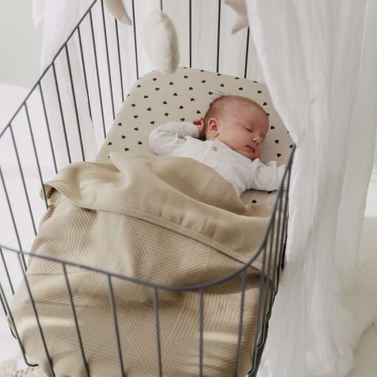 Baby slaapt in een wieg op een zandkleurig hoeslaken met zwarte hartjes, toegedekt met een beige deken.