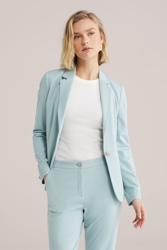Vrouw in een lichtblauwe getailleerde blazer en bijpassende broek, staand met de handen in de zakken.
