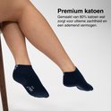 Zittende persoon met benen gekruist, draagt donkerblauwe enkelsokken met witte logo's en een donkerbruine stoel.
