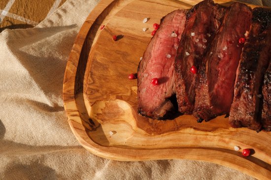 Gegrilde steak in plakjes gesneden, geserveerd op een houten plank, bestrooid met zout en rode peperkorrels.