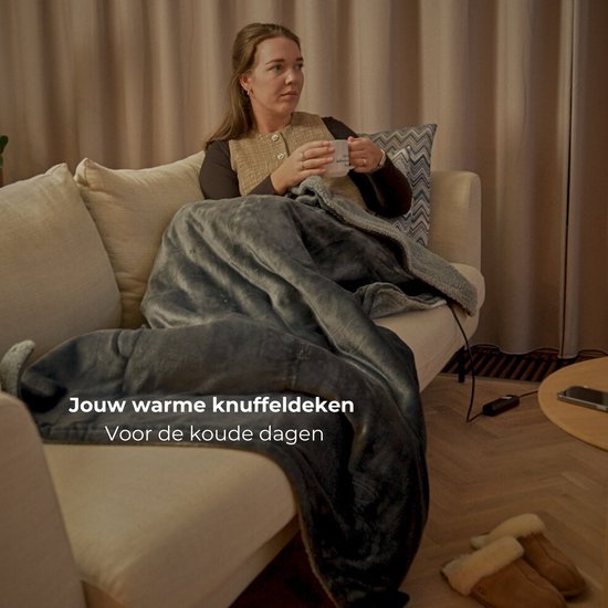 Vrouw zit op een bank, bedekt met een zachte, grijze elektrische deken, met een mok in haar handen.