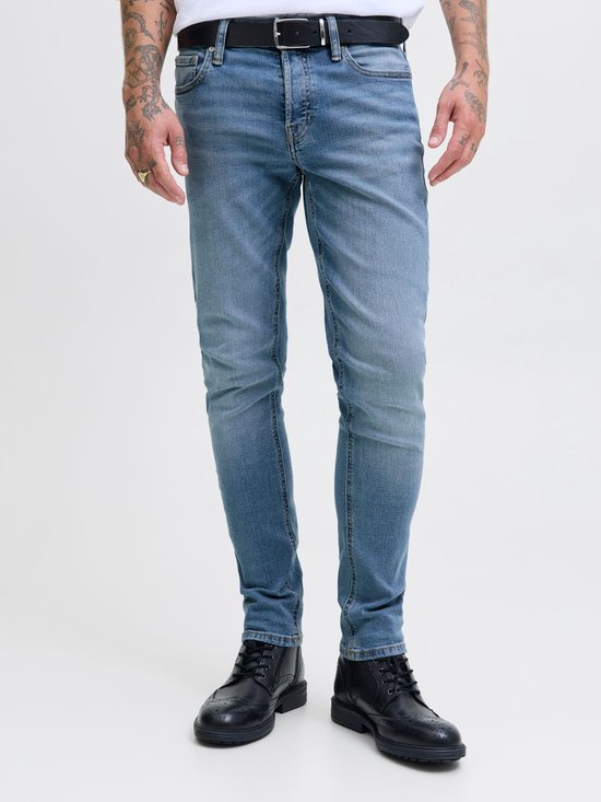 Man met tatoeages draagt een lichtblauwe skinny jeans met riem en zwarte schoenen.