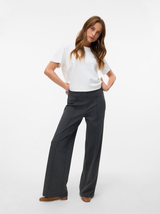 Model in een donkergrijze broek met wijde pijpen en een witte top. De broek heeft een hoge taille en verticale naden.