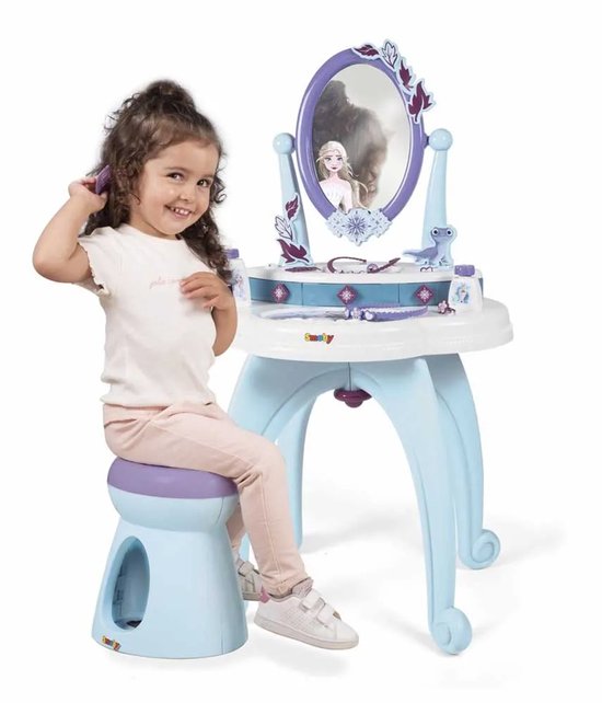 Een meisje met krullend haar zit op een blauw krukje en kamt haar haar bij een Frozen-kaptafel met spiegel.
