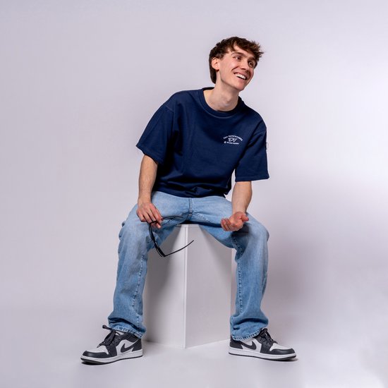 Jonge man met bruin haar, zittend op een witte kubus, lachend. Hij draagt een donkerblauwe oversized T-shirt met opdruk en een spijkerbroek.