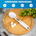 Witte lepelweegschaal met digitaal display op een houten plank. Naast de weegschaal liggen suikerklontjes en een tros druiven. Eenheden: g, oz, gn, ct.