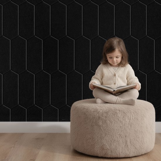 Petite fille lisant un livre sur un pouf devant un panneau mural noir à motif hexagonal.