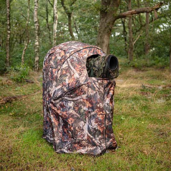 Eenpersoons camouflageschuilhut met een opening voor een camera, staand in een grasrijke bosbodem.