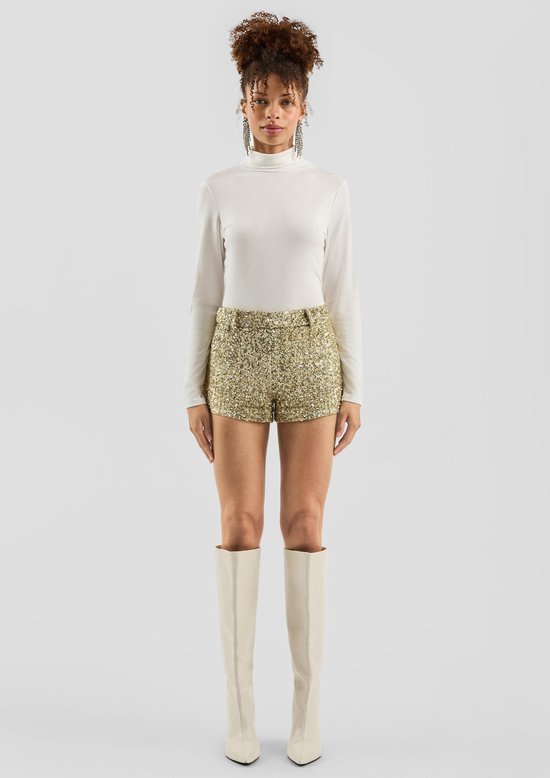Model draagt een witte coltrui, gouden pailletten korte broek, witte laarzen en grote statement oorbellen.