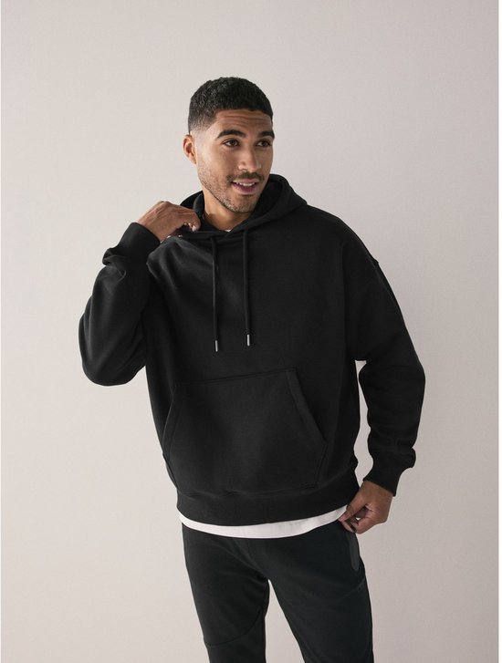Model in zwarte, oversized katoenen hoodie met capuchon, witte onderlaag en zwarte broek.