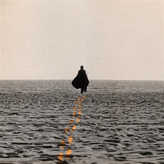 Une personne portant une cape noire marche sur une étendue de sable, laissant une traînée d'empreintes de pas orange.