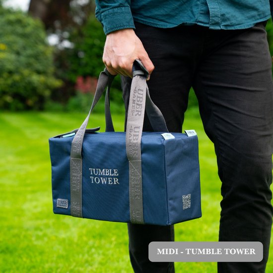 Hand met blauwe draagtas met 'TUMBLE TOWER' tekst, gedragen in een tuin.