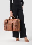 Een vrouw in een zwarte top en broek draagt een grote, bruine, leren werktas met vele buitenvakken.