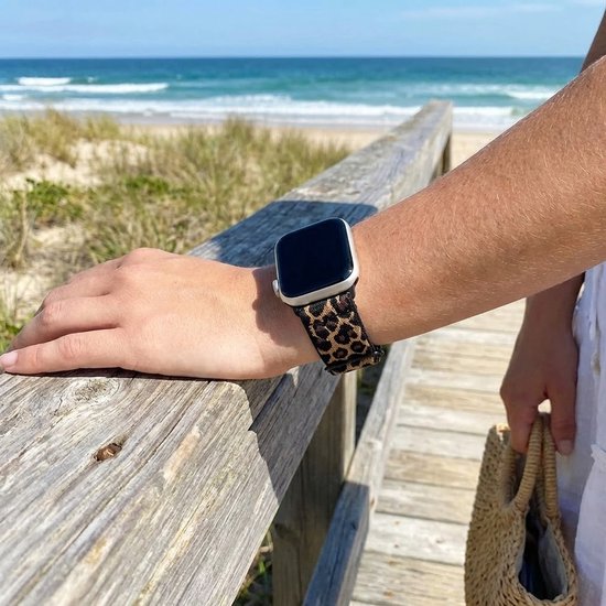 Een pols met een smartwatch en een luipaardprint horlogebandje, bij de zee op een houten promenade.