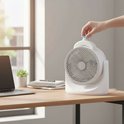 Witte tafelventilator op een bureau, hand draait aan het blauwe knopje. Laptop en plant ernaast.