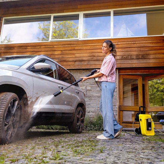 Vrouw reinigt auto met Karcher hogedrukreiniger in de tuin, met gele K4 hogedrukreiniger op de achtergrond.