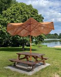 Bruine kokos parasol met houten frame boven een picknicktafel in een grasrijk park, met een meer en bomen op de achtergrond.