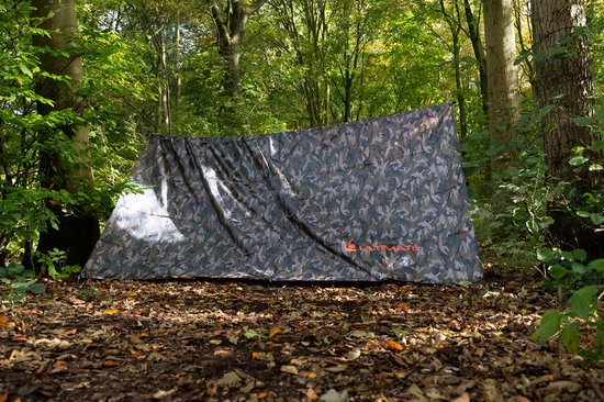 Ultimate camouflage tarp shelter, opgesteld op de bosbodem tussen bomen. Merknaam 'ULTIMATE' zichtbaar.