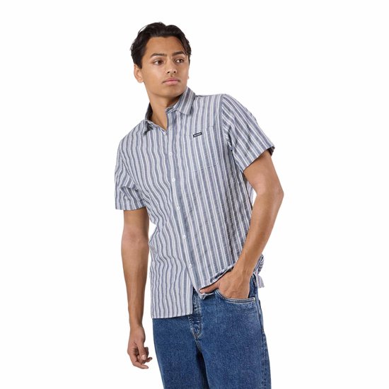 Man met de hand in zijn zak in een blauw en wit gestreept overhemd met korte mouwen en jeans.
