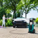Vrouw met linnen hemd en spijkerbroek reinigt zwarte auto met groene en zwarte hogedrukreiniger.