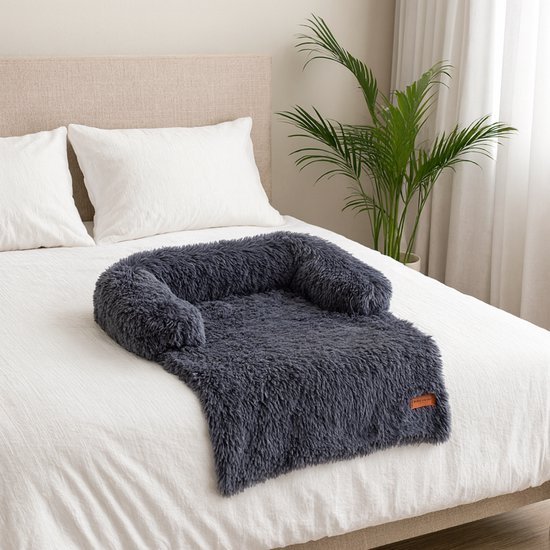 Donkergrijs, pluizig hondenkussen op een wit bed, met witte kussens en een plant.