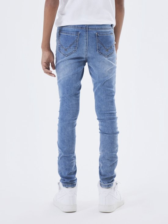 Jongensmodel in een witte t-shirt en een skinny spijkerbroek van medium blue denim, gezien vanaf de achterkant.
