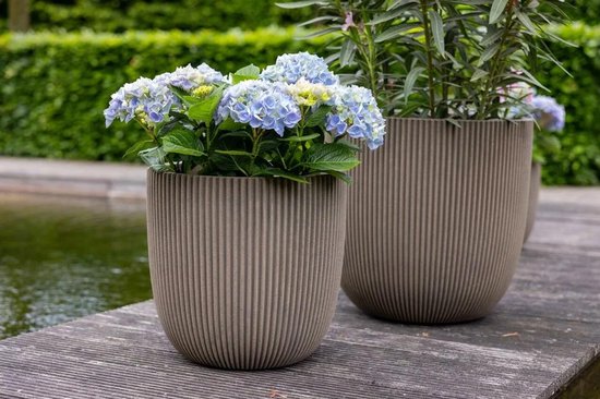 Twee taupe bloempotten met ribbelpatroon, gevuld met hortensia's en planten, op een houten vlonder bij een vijver.