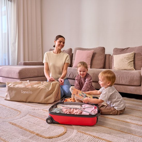 Vrouw en twee kinderen zitten op een vloerkleed bij een open rode koffer, met een beige Lionelo draagtas ernaast.